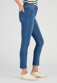 Jean skinny en denim bleu avec taille mi-haute, cinq poches et détails de couture subtils, associé à des baskets blanches.