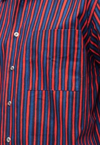 Κοντινό πλάνο ενός πουκάμισου με κάθετες ρίγες κόκκινου, μπλε και μαύρου χρώματος, με λευκά κουμπιά και τσέπη στο στήθος στην αριστερή πλευρά.