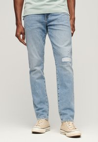 Jasnoniebieskie dżinsy o prostym kroju, z lekkim przetarciem, z łatą przy prawym kolanie i minimalnym blaknięciem.