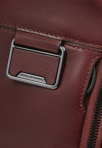 Detail einer dunkelroten Ledertasche mit silbernem Metallverschluss mit eingraviertem Logo und kontrastierendem schwarzen Reißverschluss. Glatte Textur.