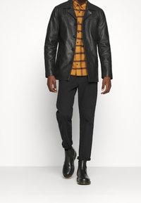 Veste en cuir noir avec fermeture à boutons, portée sur une chemise à carreaux jaune et noir en flanelle, associée à un pantalon noir et des bottines Chelsea noires.
