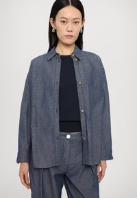 Denim srajca v temno modri barvi, s klikom na gumb, žepom na prsih in sproščenim krojem, v kombinaciji z ustreznimi hlačami.