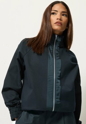 Femme aux cheveux bruns et raides portant une veste noire zippée avec des poignets élastiques, regardant légèrement sur le côté sur un fond uni.