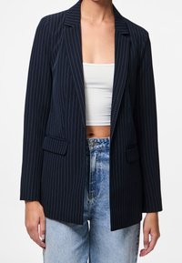 Femme portant un blazer bleu marine à fines rayures sur un crop top blanc et un jean taille haute bleu clair, debout devant un fond uni.