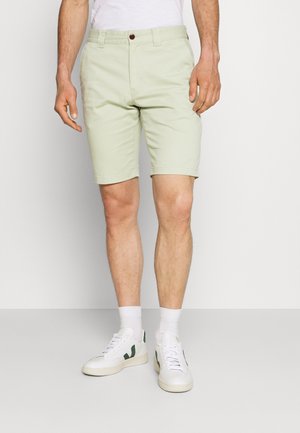 Shorts vert clair en coton avec une coupe classique, présentant une fermeture à bouton et un ourlet retroussé. Associés à des baskets blanches et des chaussettes.