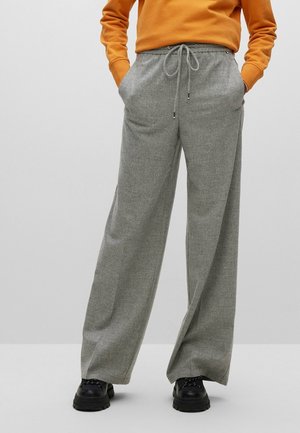 Osoba nosząca szare szerokie spodnie z sznurkiem w talii i czarne masywne buty, w połączeniu z pomarańczową bluzką z długim rękawem, ręce w kieszeniach.