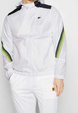 Man wearing white zip-up jacket with black and yellow patterned stripes on sleeves and matching white pants with logo patch.