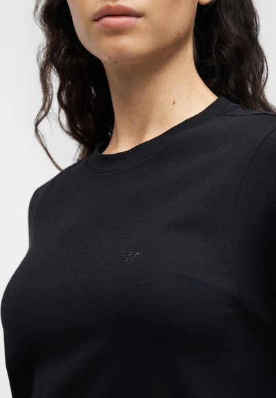 Γυναίκα φοράει απλό μαύρο t-shirt με στρογγυλή λαιμόκοψη και μικρό διακριτικό λογότυπο στο πάνω μέρος του στήθους, με σγουρά σκούρα μαλλιά.