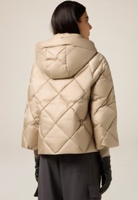 Beige gewatteerd jack met een gewatteerd ontwerp, cropped model en een capuchon. Eronder worden lange zwarte leren handschoenen en grijze broek gedragen.