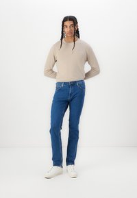 Cămășă bej cu mâneci lungi, blugi din denim albastru cu o croială clasică și adidași albi. Modelul stă pe un fundal alb simplu.