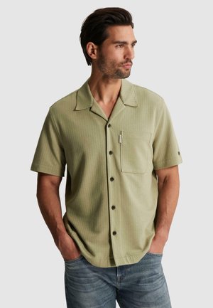 Man with dark hair and beard wearing light green short-sleeve button-up shirt and blue jeans, hands in pockets, looking right.