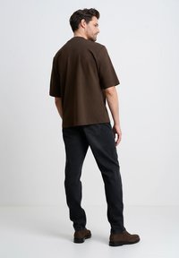 Krótka brązowa koszula z krótkim rękawem o luźnym kroju, noszona z ciemnymi dżinsami i brązowymi botkami za kostkę. Gładka faktura i minimalistyczny design.