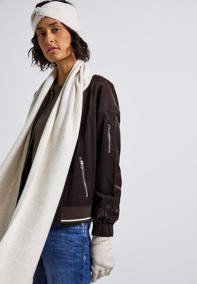 Bruine bomberjack met ritszakken, gecombineerd met een lange crèmekleurige sjaal en handschoenen. Zachte textuur; casual pasvorm met gebreide cuffs.