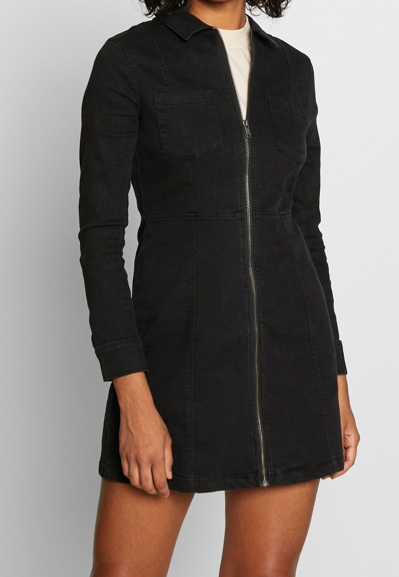 Femme portant une robe noire ajustée à manches longues avec une fermeture éclair à l'avant et des poches poitrine, debout devant un fond clair uni.