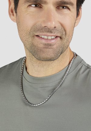 Silberne Kette mit rechteckigen Gliedern, getragen über einem grauen T-Shirt mit Rundhalsausschnitt. Glatte Textur und metallischer Glanz sind sichtbar.