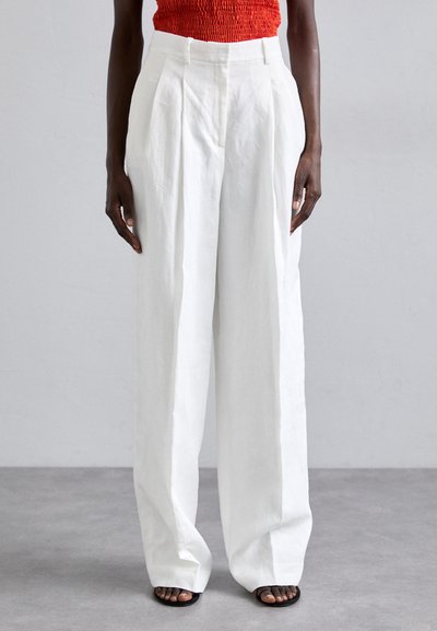 Pantalons larges en lin blanc avec des plis à l'avant, taille haute, et coupe décontractée, associés à des sandales noires à enfiler.