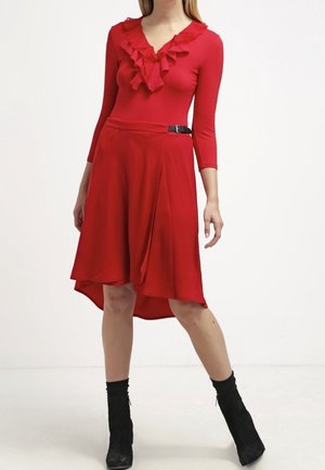 Femme portant un chemisier rouge à manches longues avec des volants et une jupe asymétrique rouge avec des bottines noires, debout sur un fond blanc.