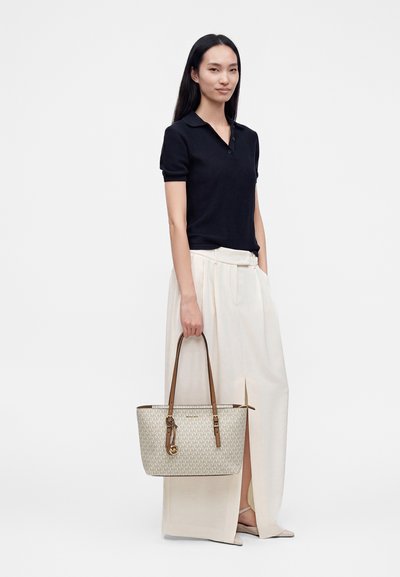 Femme aux longs cheveux noirs portant un haut noir à manches courtes et un large pantalon blanc, tenant un sac à main beige à motifs avec des anses marron.