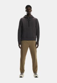 Hallikas kapuutsiga, ees koti taskuga, musta nööriga, koos khaki pükste ja heledate tossudega, seismas ühtlasel taustal.