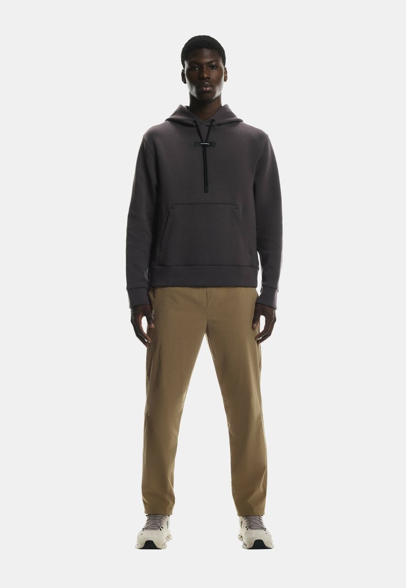 Hallikas kapuutsiga, ees koti taskuga, musta nööriga, koos khaki pükste ja heledate tossudega, seismas ühtlasel taustal.