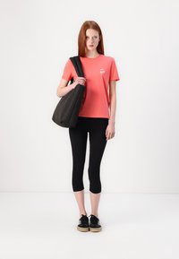 Koral t-skjorte med korte ermer og et lite grafisk motiv, svarte leggings og en svart skulderveske. Modellen har på seg svarte sko med lyse såler.