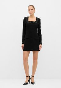 Robe noire ajustée avec des manches longues, décolleté carré, détails en perles décoratives, deux poches avant et silhouette cintrée.