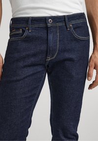 Mörkblå denimjeans med rak passform, som har orangea sömmar, en framficka och metallknäppning.