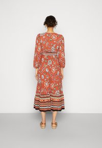 Vestido midi floral em terracota com mangas bufantes, padrões de flores azuis e brancas, e detalhe de barra listrada. Sandálias de plataforma de madeira.