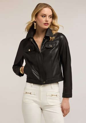 Kvinde med bølget blondt hår iført guldbelagte øreringe, sort læderjakke med lynlås foran og hvide bukser med gulddetaljer ved lynlåsen, poserer sideværts.