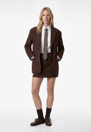 Blonde vrouw die een bruine blazer draagt, bijpassende minirok, wit overhemd, patroon stropdas, donkere sokken en bruine leren schoenen.
