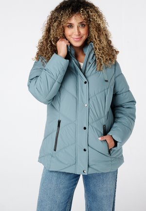 Lichtblauwe pufferjack met een ruitpatroon, hoge kraag, drukknoopjes en ritszakken aan de zijkant. Draagt over een witte top en jeans.