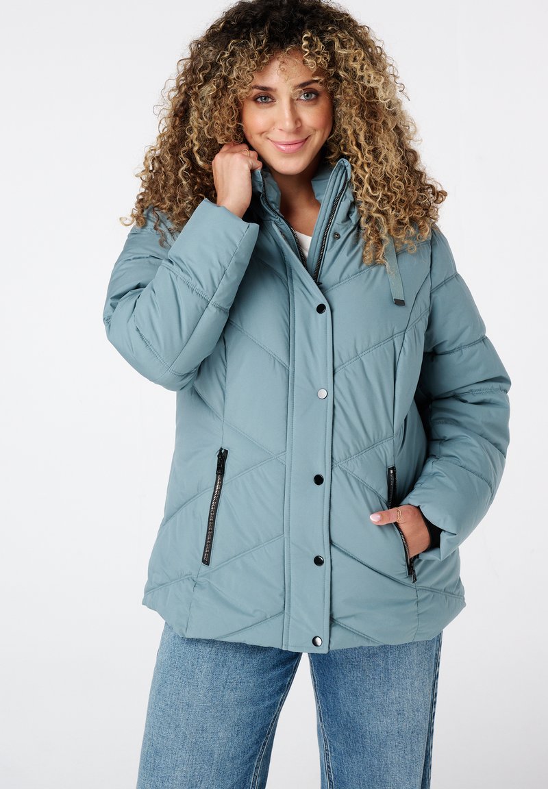 Lichtblauwe pufferjack met een ruitpatroon, hoge kraag, drukknoopjes en ritszakken aan de zijkant. Draagt over een witte top en jeans.