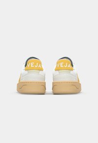 Sneakers con una toma in pelle bianca, dettagli gialli sul tallone e sul lato, e suola in gomma beige, con il nome del marchio "VEJA" sul tallone.