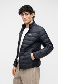 Jachetă matlasată neagră cu guler înalt, având panouri cusute și un logo EA7 pe piept. Purtați peste o cămașă albă casual.