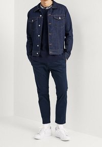 Farkkutakki nappisuljettavilla ja rintataskuilla, yhdistettynä tummansinihin housuihin ja valkoisiin lenkkitossuihin. Ribbattu pyöreä kaula-aukkosweatshirt näkyvissä.