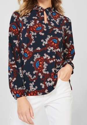Blouse marine avec un grand motif floral en rouge, bleu et blanc, présentant un décolleté en forme de clé et des manches volumineuses. Texture du tissu lisse.