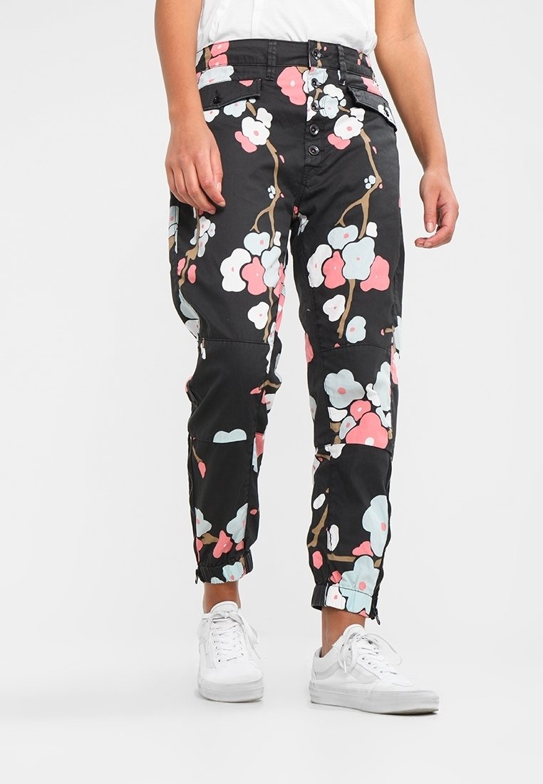 Personne portant un pantalon noir à motif floral rose et blanc, un T-shirt blanc et des baskets blanches, debout devant un fond uni clair.