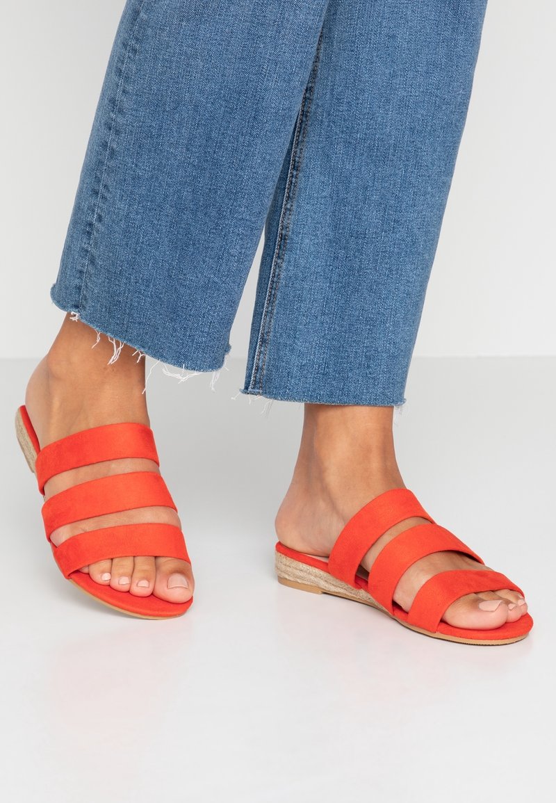 Pieds portant des sandales plates orange vif à brides, associées à un jean bleu raccourci avec des ourlets effilochés sur un sol blanc.