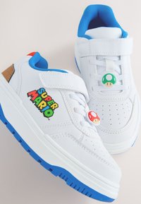 Baskets blanches avec accents bleus, dotées d'une bride en Velcro, d'un bout perforé et du logo "Super Mario". Deux détails en forme de champignon sur les lacets.