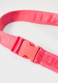 Sangle en nylon rose avec boucle en plastique, présentant une surface texturée et un lettrage en relief. Matériau solide et durable conçu pour une utilisation sécurisée.