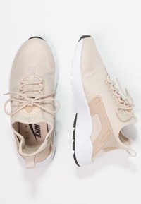 Beige Nike Air sneakers med ett ovandel i mesh, texturerade snören och en vit sula. Inkluderar en ögla på hälen och svarta gummidetaljer.