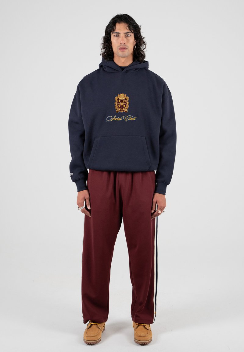 Granatowy hoodie z przeszytym złotym logo, w zestawie z bordowymi spodniami dresowymi z bocznymi paskami. Model nosi beżowe klapki.