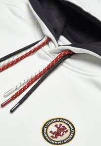 Hvid sweatshirt med en marineblå hætte, der har multicolore flettede snore og et broderet rundt emblem med et lion motiv.