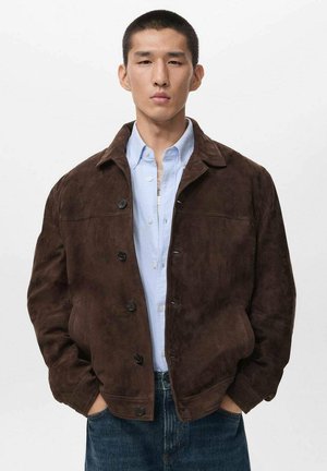 Giacca in suede marrone con chiusura a bottoni e colletto, indossata sopra una camicia azzurro chiaro. La giacca presenta una vestibilità rilassata e tasche laterali.