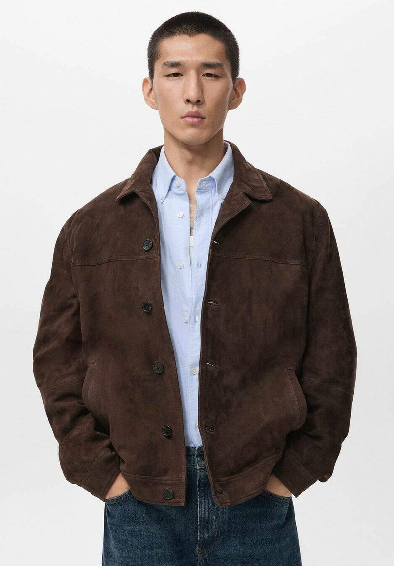 Giacca in suede marrone con chiusura a bottoni e colletto, indossata sopra una camicia azzurro chiaro. La giacca presenta una vestibilità rilassata e tasche laterali.