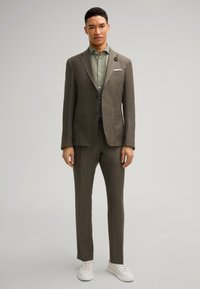 Costume marron texturé avec une coupe ajustée, associé à une chemise vert clair. Pochette blanche remarquable et baskets blanches.