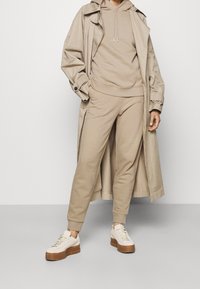 Osoba v béžové mikině s kapucí, odpovídajících joggingových kalhotách, béžovém trench coatu a bílých platformových teniskách stojící na jednobarevném pozadí.