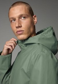 Jachetă impermeabilă de culoare verde deschis, cu glugă, având o textură netedă, guler înalt și sistem de închidere ajustabil. Design minimalist, fără logo-uri vizibile.