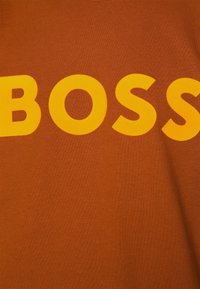 Camiseta marrón con un texto amarillo en negrita que dice "BOSS". La tela parece lisa, tiene un cuello redondo y un corte holgado.