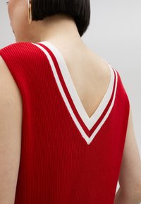 Röd ribbad ärmlös topp med en vit V-formad halsringningsdetalj. Texturerad tyg med kontrasterande ränder längs halsringningen.